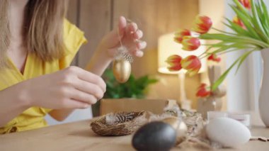 Süslü, altın Paskalya yumurtası süsü taşıyan bir kadın. Tüylü ve ipli siyah beyaz yumurta ve yuvalı ahşap masa.