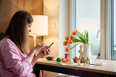 Paskalya yumurtalarını fotoğraflayan ve akıllı telefonlu el sanatları malzemeleri satan bir kadın. Tatil anları, sosyal medya ve dijital yaşam tarzını paylaşma kavramı.