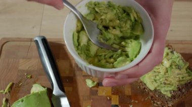 Avokado etini ahşap kesme tahtasının üzerinde çatalla kaseye sıkıştıran kişiye yaklaş. Sağlıklı kahvaltı, avokado tostu hazırlama ve vejetaryen ev yemeği kavramı.