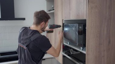 Modern mutfak dolabında vidaları olan mikrodalga fırında yapılmış yenileri tamir etmek için kablosuz tornavida kullanan bir adam. Ev aletlerinin kurulumu kavramı