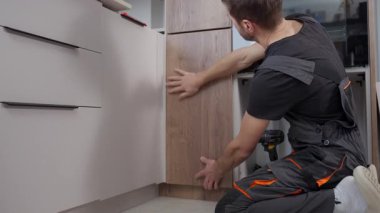 Mutfak kurulumu sırasında dolapta ahşap kapı paneli kuran iş kıyafetli adam. Tamirci evde kendi kendine mobilya monte ediyor.