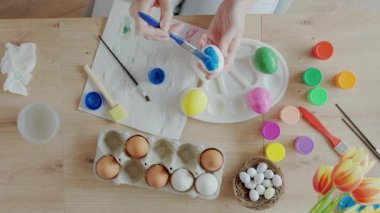 Beyaz yumurtayı fırça ve pastel boyayla boyayan kadın diğer renkli yumurtalar ve boya kavanozlarıyla aynı masaya oturuyor. Paskalya hazırlığı, yaratıcı hobi ve mevsimlik gelenek kavramı