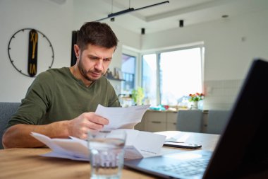 Evdeki masada oturan adam dizüstü bilgisayarla faturaları ve mali belgeleri kontrol ediyor. Kişisel finans yönetimi, bütçe ve mali stres kavramı