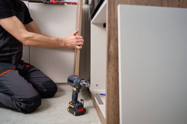 Mutfak kurulumu sırasında dolapta ahşap kapı paneli kuran iş kıyafetli adam. Tamirci evde kendi kendine mobilya monte ediyor.