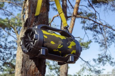 Ormandaki ağaçta asılı duran hoparlörlü kablosuz bluetooth hoparlörü. Akustik taşınabilir kamuflaj rengi, sarı kayışlı. Müzik dinlemek için aygıt.