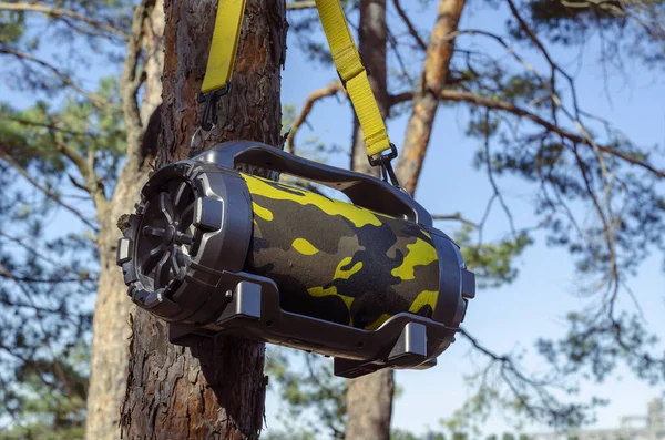 Ormandaki ağaçta asılı duran hoparlörlü kablosuz bluetooth hoparlörü. Akustik taşınabilir kamuflaj rengi, sarı kayışlı. Müzik dinlemek için aygıt.