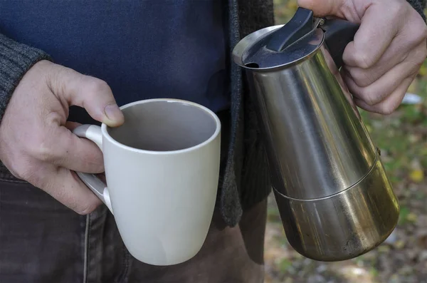 Man pours brewed coffee into cup from an outdoor geyser coffee maker. An adult man\'s hands hold ceramic cup canopy and metal coffee maker with coffee.