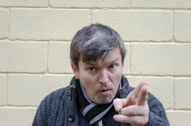 Öfkeli, el kol hareketi yapan bir adamın portresi. Orta yaşlı grimsi bir adam, sarı arka planda kızgın bir yüzle kameraya bakıyor..