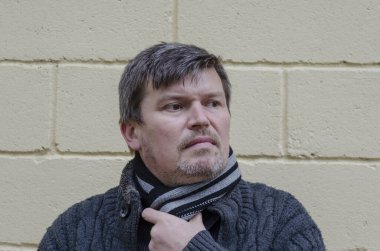 Beyaz saçlı, kendine güvenen bir adamın portresi sarı duvara bakıyor. Gri kazak ve eşarp giyen orta yaşlı adam düşünceli bir şekilde silinecek..