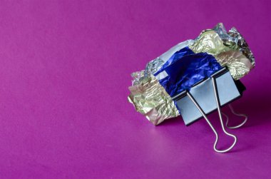 Ezilmiş şeker ambalajları ve mor arka planda metal siyah cilt klipsi. Bağlayıcı kıskaçlar rastgele çikolata ambalajlarını yapıştırıyor. Soyut yaratıcı arkaplan.