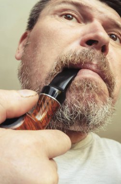 Ağzında sigara piposu olan sakallı bir adamın alışılmadık portresi. Gri sakallı ve funda kökü tüten piposu olan yetişkin bir adam. Kameranın önünde suratlar ekşiyor.