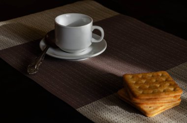 Bir yığın tuzlu kare kraker, kaşık ve fincan tabağı kafede masanın üzerinde olan beyaz kahve fincanı. Sabah kahvesi. İnsan yok. Seçici odak.
