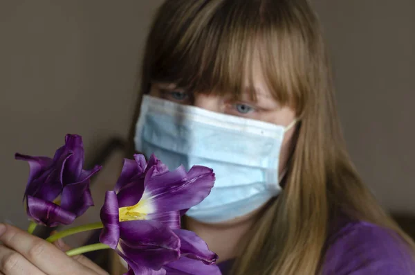 Woman in a median mask and tulips. Frontal shot of a woman with allergy wearing a medical mask and looking at colors. A woman with her hair looks at a bouquet of tulips. Healthcare and medicine. Focus on lilac flowers.