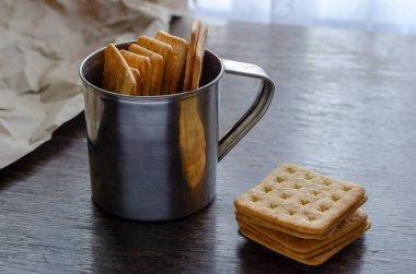 Kare tuzlu krakerler ahşap masada metal bardakta. Gevrek atıştırmalıkların bir kısmı boş paslanmaz çelik kupanın içinde. Seçici odak.