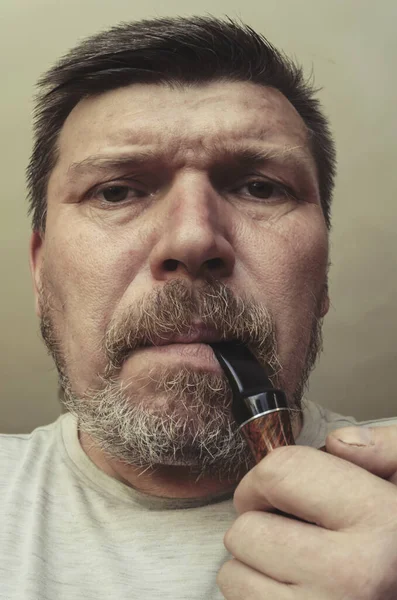 Unusual portrait of a bearded man with a smoking pipe in his mouth. A grown man with gray stubble and a heather-root smoking pipe. Grimaces in front of the camera