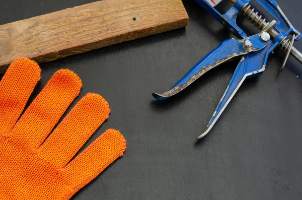 Orange work glove, sealant gun and wood rail on black table. View from above at an angle. Selective focus.