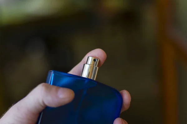 Close-Up Of Hand Holding Perfume Against dark green Background. Man\'s hand with blue glass bottle of perfume.