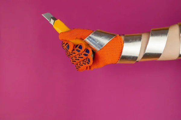 Hand wrapped with aluminum duct tape for padding. Man's hand with a yellow knife with interchangeable blades on a pink background.