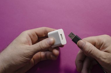 DC adaptörü ve USB veri kablosu erkeklerin elinde. Erkek, siyah kabloyu ve beyaz Amerikan duvar adaptörünü birbirine bağlıyor. Üst görünüm, pembe arkaplan.