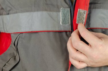 İş için özel koruyucu giysiler. Adamın eli, gri bir iş ceketinin düğmelerini kırmızı eklerle ilikliyor. Yakın çekim, seçici odaklanma.