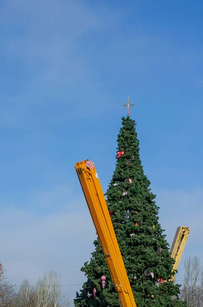 市内の主要クリスマスツリーを解体する 黄色のブーム青い空の背景に装飾が施された2つの移動式クレーンと背の高いクリスマスツリー 人はいない — ストック写真