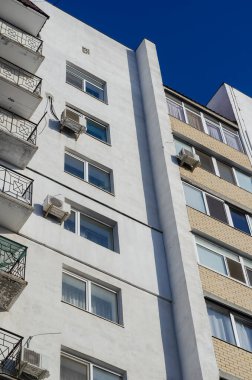 Mavi gökyüzüne karşı apartmanın zemin manzarası. Güneşli bir günde balkonları ve kliması olan beyaz bir bina. Emlak, kiralama..