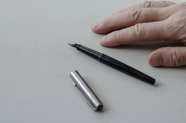 Man's right hand, black fountain pen, and metal pen cap lie on the light-colored table. Close-up. Selective focus.