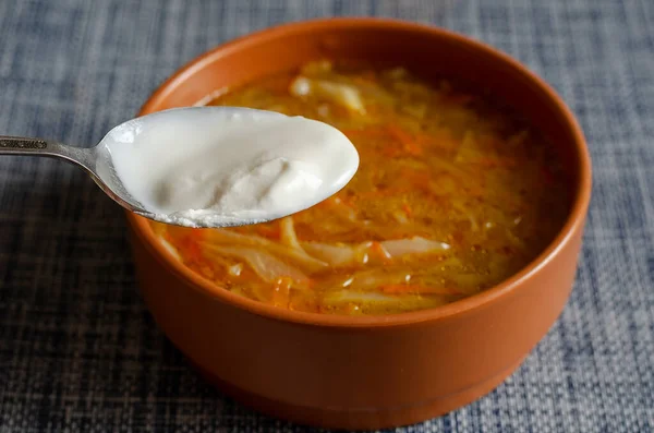 A spoon with sour cream and cabbage soup in a brown earthenware bowl on a blue tablecloth. A serving of ready-to-eat sauerkraut soup. Close-up, selective focus.