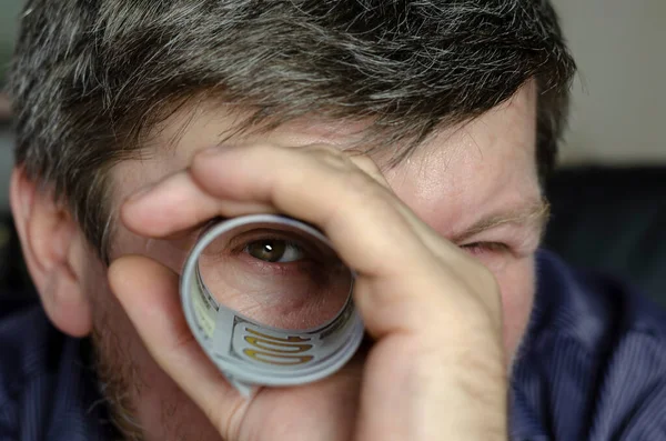 The person looks through the rolled money with one eye. A middle-aged man with gray hair looks through a tube of dollars.