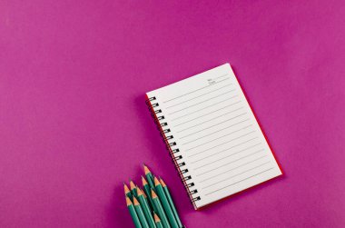 Pembe arka planda yaylı defter ve kalemleri aç. Beyaz not defteri ve yeşil kalemler. Yukarıdan açılı görüş. Seçici odak.