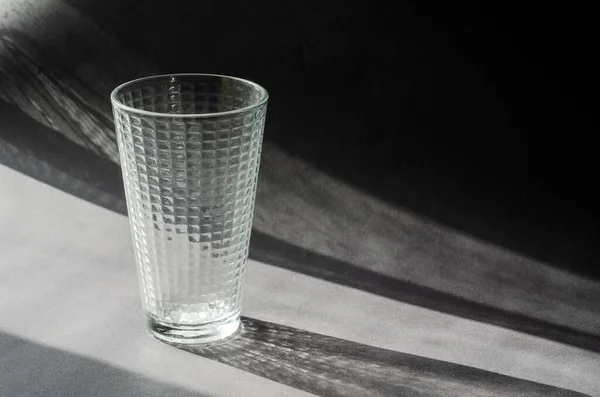 Empty glass beaker on a gray surface with shadows. Transparent glass with a checkered pattern. Abstract sun shadows on gray. Copy space, selective focus.