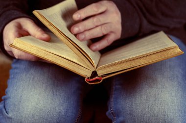 Adamın ayaklarının dibinde açık bir kitap yatıyor. Mavi kotlu ve siyah gömlekli bir adam sayfaları çeviriyor. Göz hizasında ateş ediliyor. Yakın çekim, seçici odaklanma.