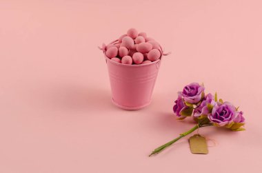 Pembe arka planda Dragee, kova ve çiçeklerle minimalist bir hayat. Şeker dolu metal bir kova. Etiketli bir buket yapay gül. Yakın çekim, seçici odaklanma.