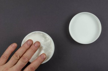Gri arka planda kozmetik kremi olan el ve konteynır. Bir adamın eli ve kapaklı yuvarlak beyaz bir krema kavanozu..