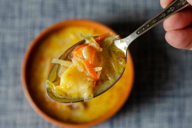 El, lahana çorbası ile metal bir kaşık tutuyor. Arka planda çorba olan kahverengi bir toprak kap. Yakın plan, seçici odaklanma. Üst görünüm.