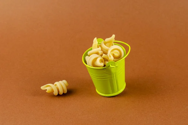 Green metal bucket and dry insalatonde pasta on a brown background. The full Small bucket of pasta is tilted. Close-up, selective focus