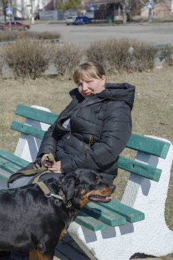 Sokak bankında bir kadın ve tasmalı bir evcil hayvan. Yetişkin bir kadın ve Rottweiler dinleniyor. Uzun saçlı, orta yaşlı bir kadın. Bahar güneşli gün.
