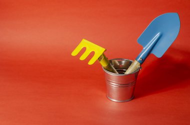 Sarı çocuk tırmığı ve mavi bahçe kepçesi kırmızı arka planda küçük bir kovada. Bahçe aletleriyle minimalist kompozisyon. Yakın plan. Seçici odak.