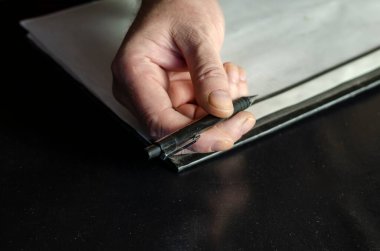 Memurun elleri yakın planda siyah bir masanın üzerinde bir kalem, bir dosya klasörü tutuyor. Yeşil kamuflaj üniformalı orta yaşlı bir adam. İçeride. Seçici Odaklanma. Alçak tuş.