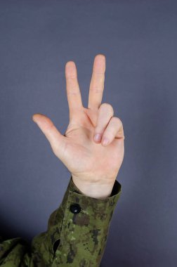 El kamuflajı, parmaklarının gri arka planda olduğunu gösteriyor. Üç parmak kaldırıldı. Orduda üç numaranın ataması. Ordu jest dili.