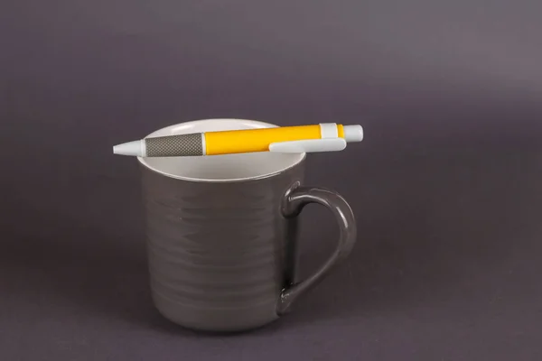 灰色の背景に灰色の縞模様のマグカップと黄色のペン プラスチックペン付きの空の新しいセラミックマグカップが上にあります ミニマリズム — ストック写真