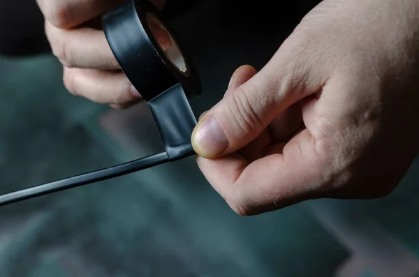 Bir adam siyah bir elektrik telini yalıtıyor. Elektrik işi için bir rulo siyah koli bandı tutan erkeklerin yakın çekimi. Yetişkin bir erkek, beyaz. Odanın içinde. Seçici odak.