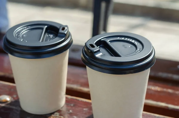 Two paper cups with a lid on a park bench. Close-up of eco-friendly disposable hot drinks takeaway tableware. Daytime. Outdoors.