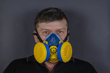 Gri arka planda endüstriyel solunum cihazındaki bir adamın portresi. Kısa saçlı, mavi ve sarı solunum cihazı takan yetişkin bir erkek. Değiştirilebilir filtreli yarı maske. İçeride. Güvenlik önce gelir.