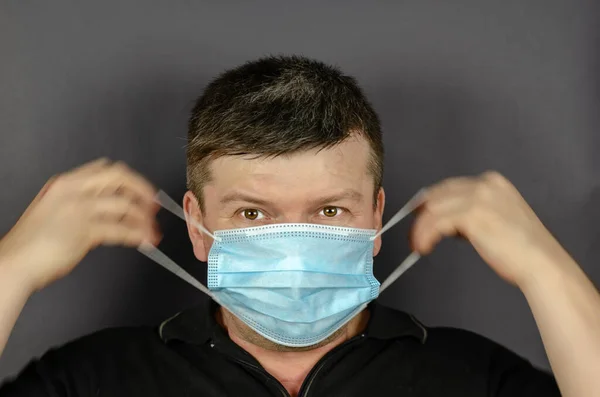An adult man puts on a medical mask on a gray background. A middle-aged male with brown eyes and short graying hair. Blurry hand movement. New normal lifestyle. Healthcare and medicine.