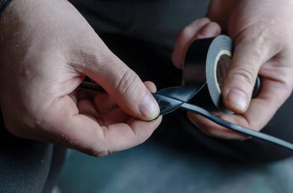 Bir adam siyah bir elektrik telini yalıtıyor. Elektrik işi için bir rulo siyah koli bandı tutan erkeklerin yakın çekimi. Yetişkin bir erkek, beyaz. Odanın içinde. Seçici odak.
