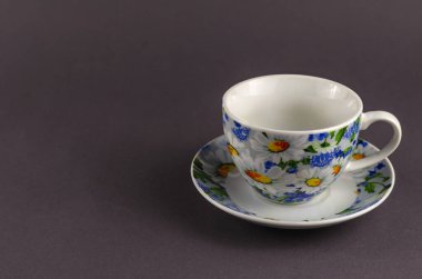 Beyaz fincanda gri arka planda boyanmış çiçekler var. Çay için seramik bardak ve çay tabağı ya da parlak mavi ve sarı renklerle süslenmiş kahve. Tabaklarda beyaz papatyalar.