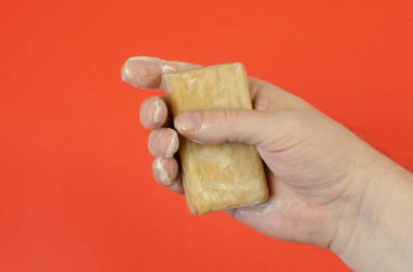 Kırmızı arka planda el ve ıslak çamaşır sabunu. Adamın elinde dikdörtgen bir kalıp kahverengi sabun var. Avuç içi sabunlu köpükle kaplı..