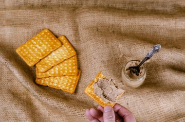 Krakeri karaciğer ezmesiyle tutan bir elin yüksek açılı görüntüsü. Üzerine et bulaşmış yarım bir aperatif tutan adam. Arka planda kaşık olan krakerler ve kavanozlar. Seçici odaklanma. Ev yapımı yemek..