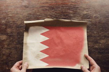 Eski kağıt üzerinde beyaz ve kırmızı bir Bachrane bayrağı. Bir adamın elleri parşömen kağıdını kahverengi bir tahta masaya bastırıyor. Yukarıdan görüntüle